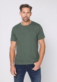 Zelené tričko s krátkým rukávem vyrobené z bavlněné látky, s kulatým výstřihem a malou kapsou na levé straně hrudi. Hladká texturu.