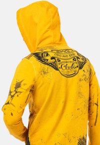Haut à capuche jaune avec un imprimé graphique mettant en avant le logo d'un club de moto, des accents noirs et un aspect texturé et usé.