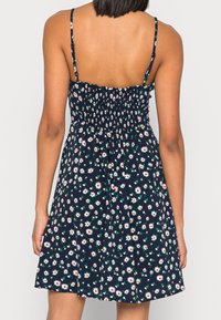 Žena v tmavě modrých šatech bez rukávů s bílým květinovým potiskem sedmikrásek a nařaseným zadním dílem, viděná zpoza.