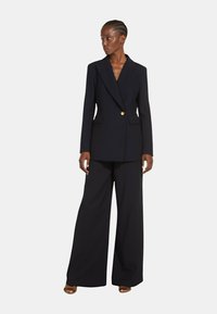 Blazer blu navy con un singolo bottone dorato, rever a punta e tasche laterali, abbinato a pantaloni a gamba larga in tessuto coordinato.