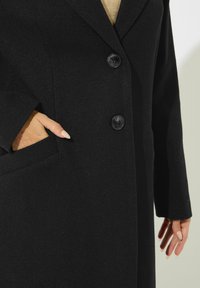 Cappotto in lana nera con design sartoriale, due bottoni neri e una tasca laterale. Il tessuto ha una finitura strutturata, che offre calore e stile.