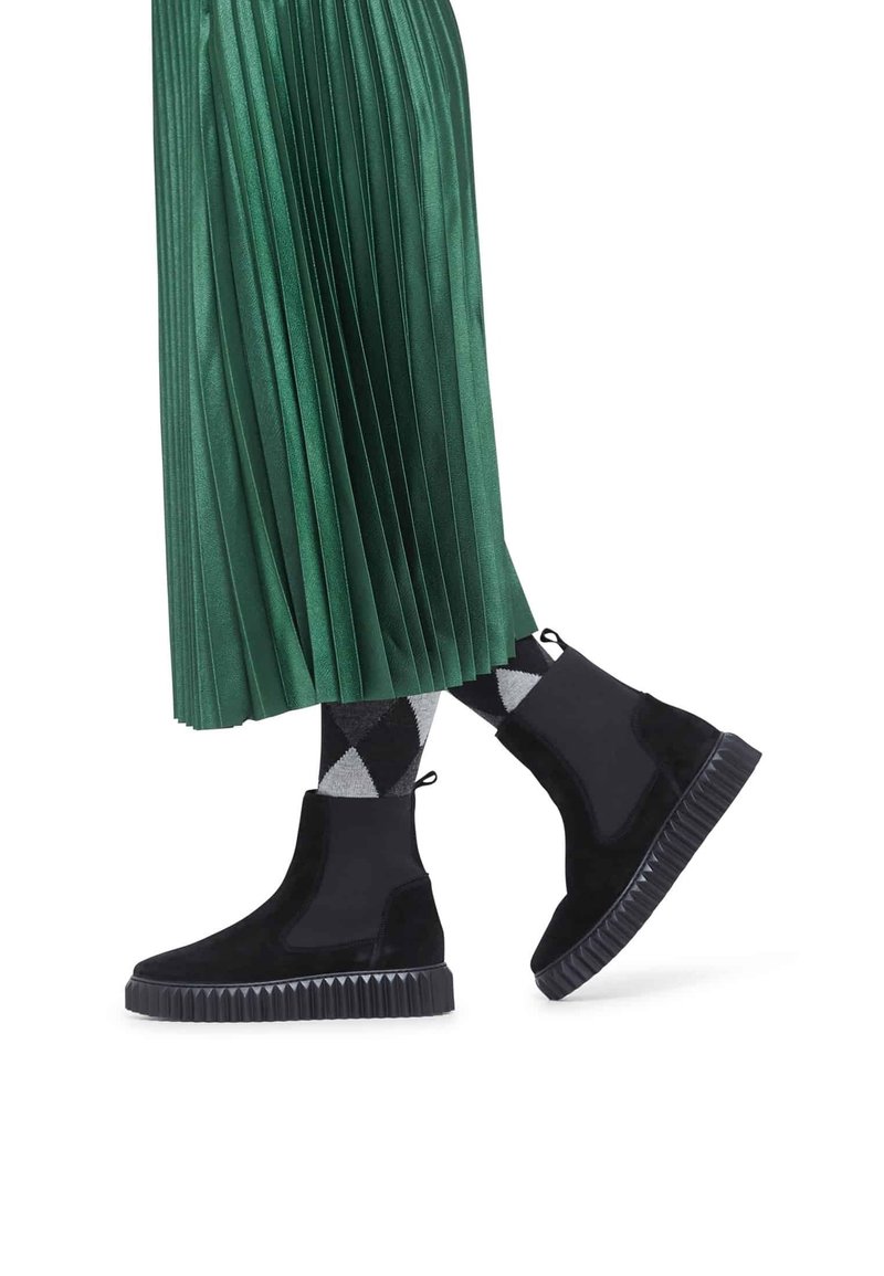 Jambes en bottines à plateforme noires avec des chaussettes à losanges noires et grises, portant une longue jupe plissée verte, marchant sur un fond blanc.