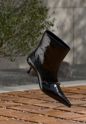 Bottine noire brillante à bout pointu avec petit talon flottant au-dessus d'un sol en bois et en pierre à l'extérieur.