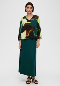 Geprinte top met gele, groene en bruine abstracte patronen, gecombineerd met een effen donkergroene maxi rok. Sandalen maken de outfit compleet.