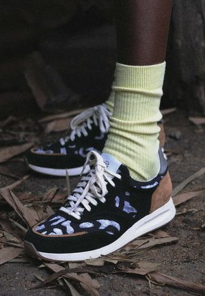 Personne portant des baskets à motifs noirs et blancs avec des accents sable et des chaussettes côtelées jaune clair, debout sur un sol couvert de feuilles sèches.
