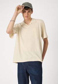 Polo color beige con collar, mangas cortas y un bolsillo en el pecho, combinado con jeans de color azul oscuro. El modelo lleva una gorra gris.