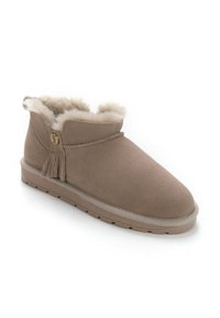 Beige nahkahousut nilkkojen ylle, joissa on pehmeä fleece-sisus, koristeellinen tupsu ja kumipohja. Pyöristetyt varpaat ja ommellut yksityiskohdat.