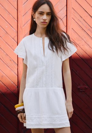 Jeune femme aux longs cheveux foncés portant une robe blanche à manches courtes et des bracelets colorés, debout devant une porte en bois rouge.