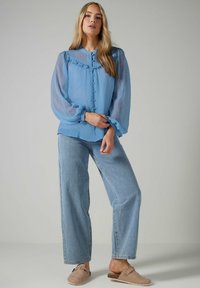 Blusa ligera azul claro, transparente, con patrón de lunares, cuello con volantes y cierre de botones. Combinada con jeans de talle alto azul claro y zapatos tipo slip-on.