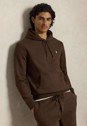 Jeune homme aux cheveux bouclés portant un sweat à capuche marron et un pantalon de jogging assorti, debout les mains dans les poches devant un mur uni.