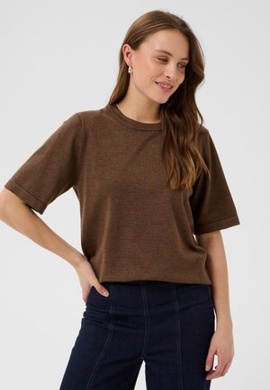 Femme aux longs cheveux bruns portant un haut marron à manches courtes et un jean foncé, se tient debout, une main sur la hanche, souriant légèrement.