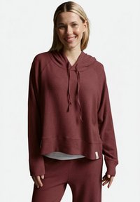 Lächelnde Frau in einem kastanienbraunen Kapuzenpullover mit langen Ärmeln und Kordelzügen sowie passenden Hosen, die vor einem schlichten weißen Hintergrund steht.