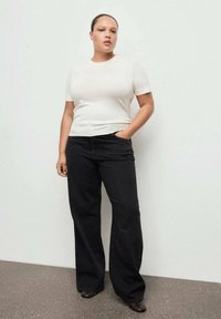 Bela oprijeta majica s kratkimi rokavi v kombinaciji z visokim pasom in črnimi širokimi kavbojkami. Model stoji na svetli podlagi.