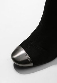Botte en daim noir avec un embout métallique brillant gravé de "Calvin Klein" sur fond blanc.