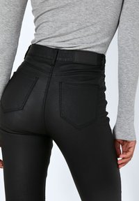 Zwarte high-waisted skinny jeans met een gladde textuur, voorzien van twee achterzakken en gestikte details, gecombineerd met een grijze lange mouwtop.