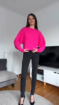 Maglione oversize di un rosa acceso con maniche lunghe a palloncino, abbinato a pantaloni neri aderenti e décolleté nere. Ambientazione interna.
