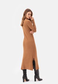 Długa, prążkowana sukienka dzianinowa w kolorze brązowym z rozcięciem z tyłu i dopasowanymi rękawami, w połączeniu z wysokimi czarnymi botkami na obcasie. Gładka struktura i dopasowany krój.