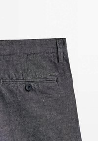 Poche arrière d’un pantalon gris foncé texturé avec fermeture par bouton et une petite étiquette de marque rectangulaire sur le côté droit.