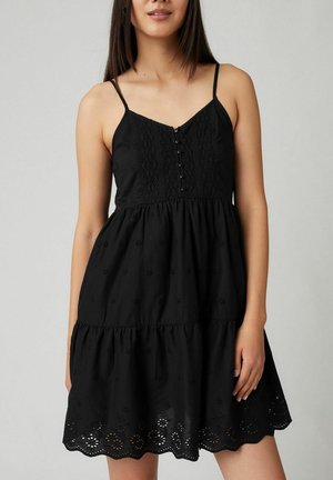 Woman wearing sleeveless black dress with lace details, buttons on bodice, and scalloped hem with eyelet floral embroidery.