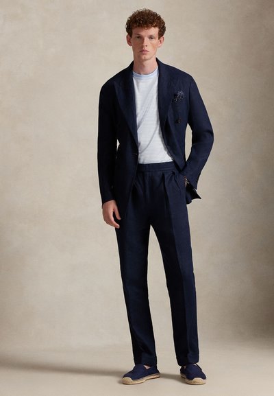 Τζιν κοστούμι σε ναυτικό μπλε με διπλό πέτο, παντελόνια με πιέτες και ριγέ λευκό t-shirt. Συμπληρώνεται με μαντηλάκι τσέπης με σχέδιο και εσπαντρίγιες.