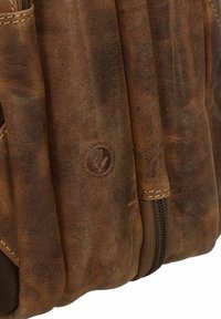 Sac à dos en cuir marron avec une finition texturée, des coutures visibles et un tampon de logo. Il dispose d'une fermeture éclair sur le côté.