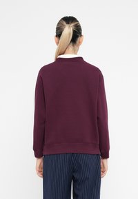 Oseba z blond konjskim repom, oblečena v maroon pulover in hlače s temno modrimi črtami, prikazana od zadaj na beli podlagi.