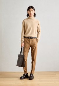 Bež turtleneck pulover in svetlosive hlače, dopolnjeni z temno torbo in rjavimi vezalkami, stoječi na leseni tleh.