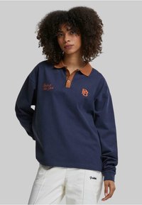 Marineblå langermet polo med brun krage, med oransje broderi som sier "Berlin til New York" og en logo på brystet.