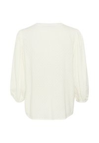 Blusa blanca con patrón de lunares texturizados, cuello redondo y mangas abullonadas. Los dobladillos tienen una ligera curvatura para un ajuste relajado.