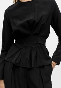 Haut peplum noir en tissu lisse, avec des manches longues, un col rond et un design plissé à la taille. Coupe simple et élégante.