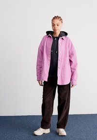 Ružová kordová bunda so striebornými gombíkmi cez čiernu mikinu, doplnená hnedými kordovými nohavicami a béžovými teniskami, na neutrálnom pozadí.
