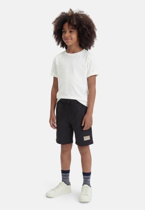 Kind steht und trägt ein weißes T-Shirt, schwarze Shorts mit einem Aufnäher, gemusterte marineblaue Socken und weiße Turnschuhe vor weißem Hintergrund.