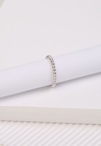 Silberner Ring mit einem Perlenmuster und glatter Oberfläche, der auf einem weißen zylindrischen Objekt vor einem weißen Hintergrund platziert ist.