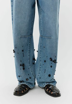 Pantaloni din denim albastru deschis cu picioare largi, decorați cu broderii florale negre. Asortați cu sandale negre împletite, cu talpă groasă și design deschis.