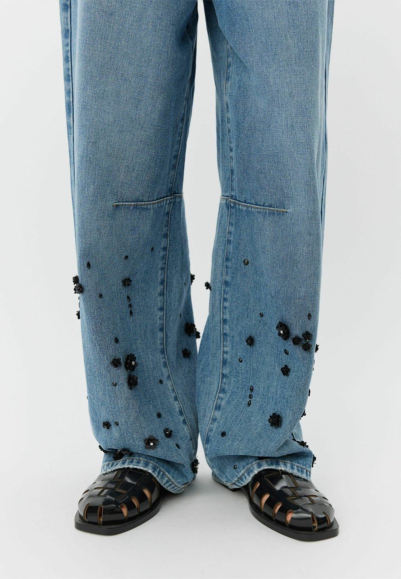 Svetlomodré džínsové nohavice s širokými nohavicami a čiernymi kvetinovými ozdobami. Kombinované s čiernymi pletenými sandálmi s hrubou podrážkou a otvoreným dizajnom.