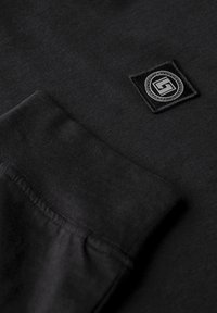 Black sweatshirt featuring a square patch with a logo and circular border. The fabric is smooth with visible stitching details.