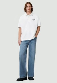 Jovem em pé com as mãos parcialmente nos bolsos, vestindo uma camisola polo branca da AllSaints, calças largas de ganga azul-claro e sapatos pretos.