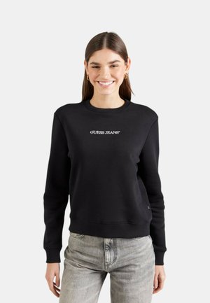 Giovane donna sorridente, indossa una felpa nera con la scritta "GUESS JEANS" e jeans grigio chiaro, in piedi contro uno sfondo chiaro uniforme.