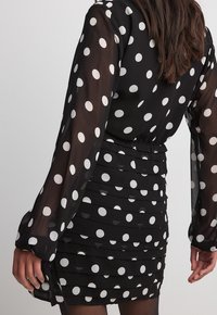Vestido negro con lunares, mangas largas de gasa, textura fruncida y silueta ajustada. Presenta patrones circulares blancos por toda la prenda.