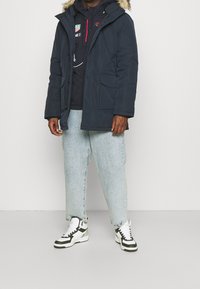 Granatowa parka z kapturem obszytym futrem, zapinana na zamek i guziki, z dwoma przednimi kieszeniami, noszona na ciemnej bluzie i jasnoniebieskich dżinsach, w połączeniu z trampkami.