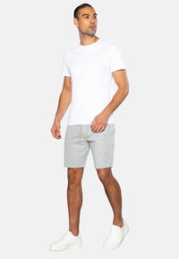 Hvid bomulds t-shirt, grå shorts med gule snore, og hvide sneakers. Modellen står i en neutral stilling foran en ensfarvet baggrund.