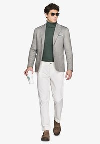 Grå blazer over en grøn turtleneck, hvide bukser og brune loafers. Holder en hvid rejsekop med et grønt logo. Solbriller.