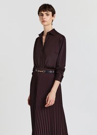 Woman wearing a dark brown satin button-up shirt tucked into a matching pleated skirt with a black belt against a plain background.