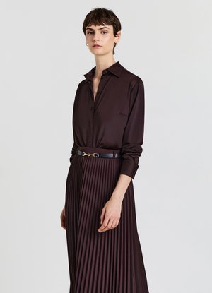 Donna che indossa una camicia in satin marrone scuro con bottoni, infilata in una gonna plissettata dello stesso colore, con una cintura nera, su uno sfondo semplice.