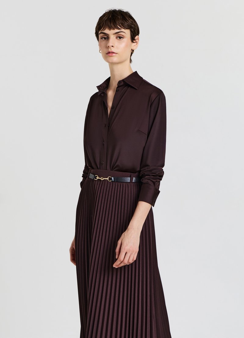 Woman wearing a dark brown satin button-up shirt tucked into a matching pleated skirt with a black belt against a plain background.