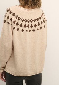 Beige stickad tröja med ett brunt geometriskt mönster över axlarna, med en avslappnad passform och ribbad nederkant. Mjuk textur, långa ärmar.