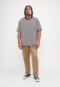 Kortermet svart-hvitt mønstret skjorte, avslappet passform, kombinert med beige bukser og svarte sneakers, stående på en nøytral bakgrunn.