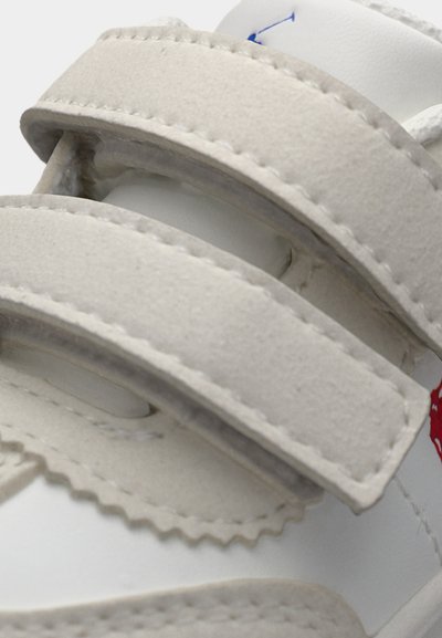 Κοντινό πλάνο άσπρου αθλητικού παπουτσιού με δύο μπεζ λουριά βέλκρο από σουέντ και ορατές λεπτομέρειες ραφής στο επάνω μέρος.