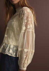 Blusa in pizzo color crema con maniche a sbuffo trasparenti, ricami floreali e orlo con volant. Cuciture dettagliate e motivi testurizzati arricchiscono il design.