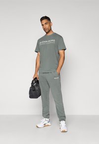 Γκρίζα βαμβακερή μπλούζα και φόρμες με κείμενο "BJÖRN BORG", συνδυασμένα με μαύρη τσάντα γυμναστηρίου και λευκά αθλητικά παπούτσια με ταμπέλα καφέ.